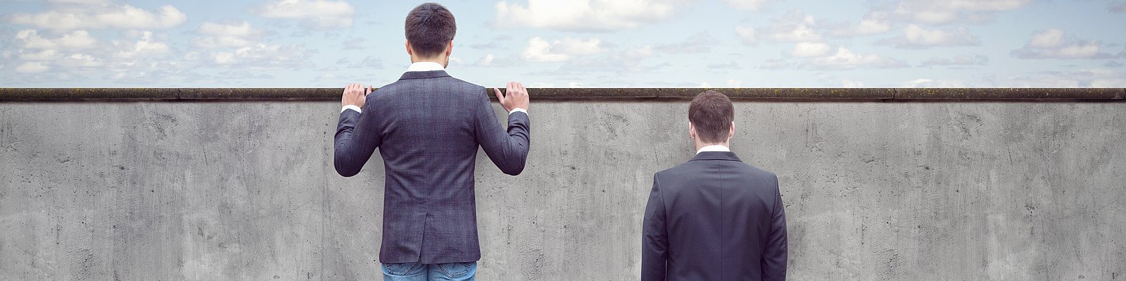 Zwei Männer stehen vor einer Mauer, einer davon auf einem Bücherstapel schaut über die Mauer hinweg