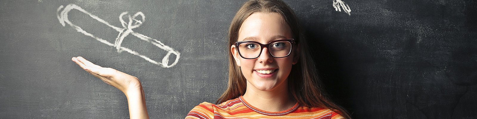 Junge Frau vor einer Schiefertafel mit angezeichneter Diplomrolle über der Hand und Graduiertenhut über dem Kopf AdobeStock_96461795.jpeg