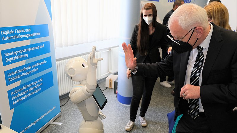 Mann mit Maske hebt Hand und gegenüber hebt ein Roboter auch die Hand zum Gruße